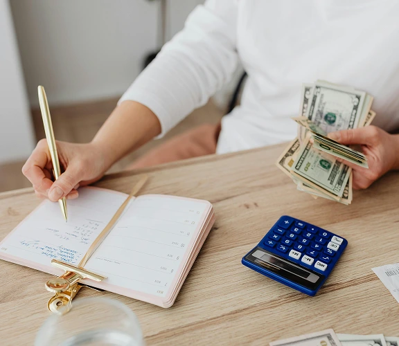 A woman writing in her notepad with cash in the other hand and a calculator at the desk